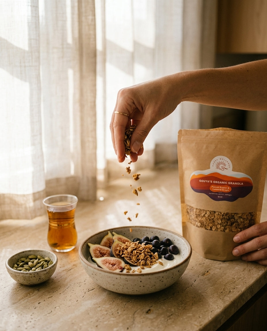 Hand sprinkling granola over a fruit and yogurt bowl by a window, with the Gouya granola bag nearby