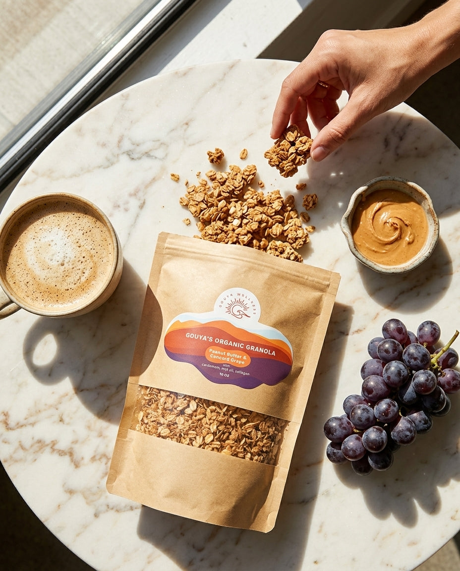 Hand taking granola from a marble table beside the bag, a coffee, peanut butter and grapes